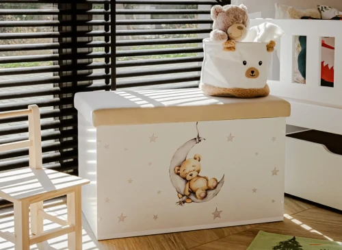 Hvid legetøjskasse med bamse-motiv, lille træstol og bamse-opbevaringspose ved vinduet.