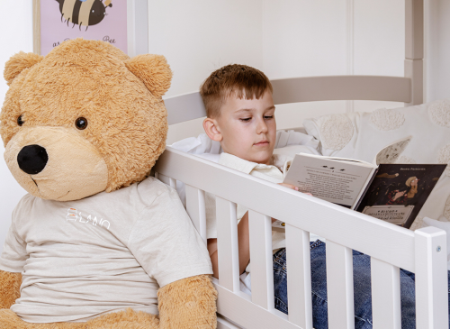 Dreng læser en bog i en hvid seng ved siden af en stor brun bamse, rolig læsestund i børneværelset.