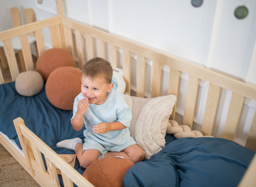 Glad barn sidder i en hus-tremmeseng i naturtræ med blå dyne og puder.