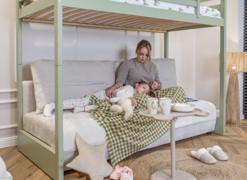 Mor og sovende barn på sofa under grøn køjeseng med tæppe og drikkevarer. Afslappet stemning i børneværelset.