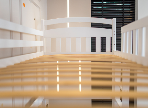 Hvid sengeramme i træ med lameller set fra siden i et lyst børneværelse.