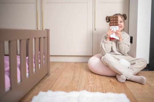 En pige sidder på gulvet og dækker sit ansigt med en plade chokolade. Til venstre ses en del af sengen i mælkechokolade-farve.